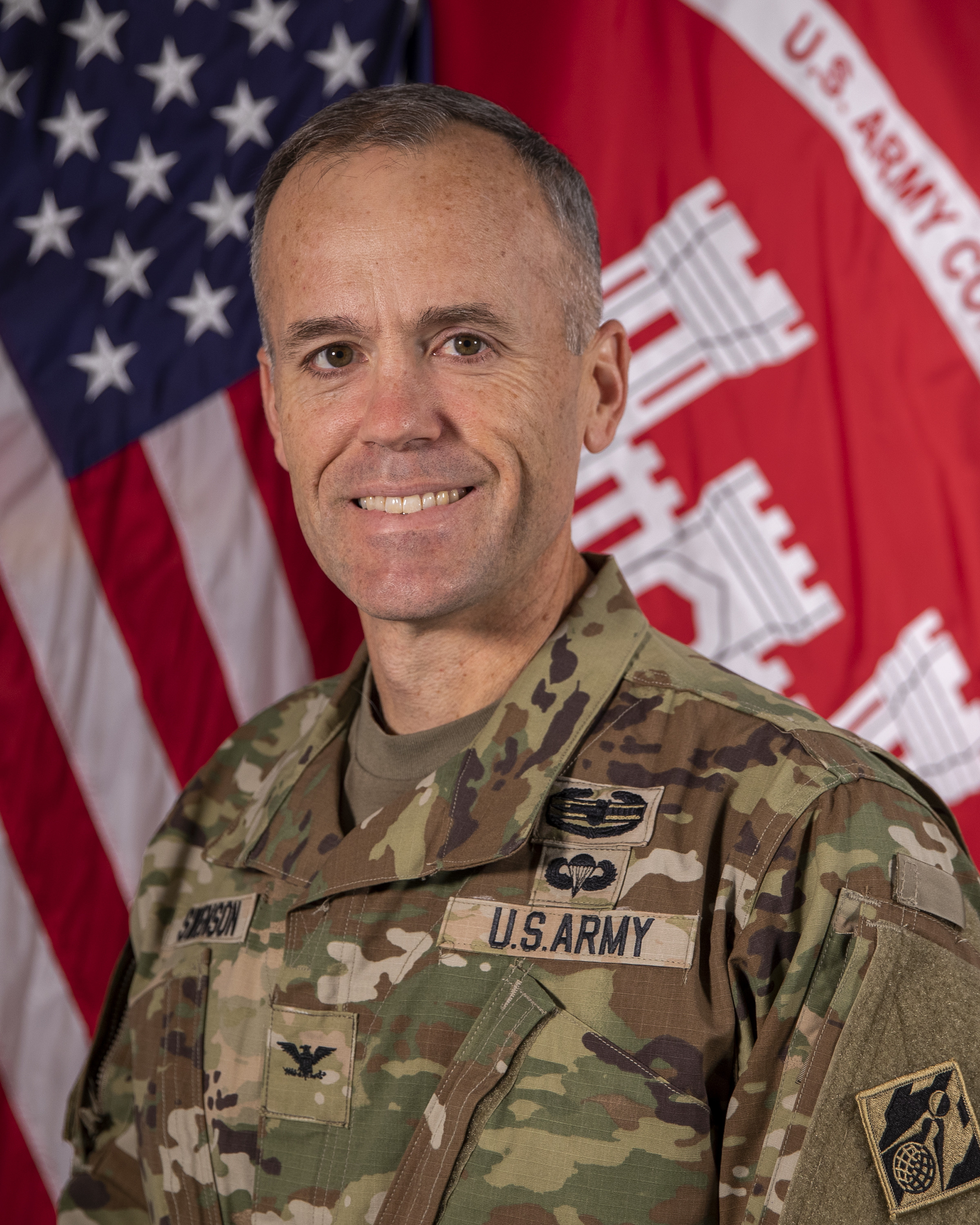 A man in a military uniform stands in front of two flags