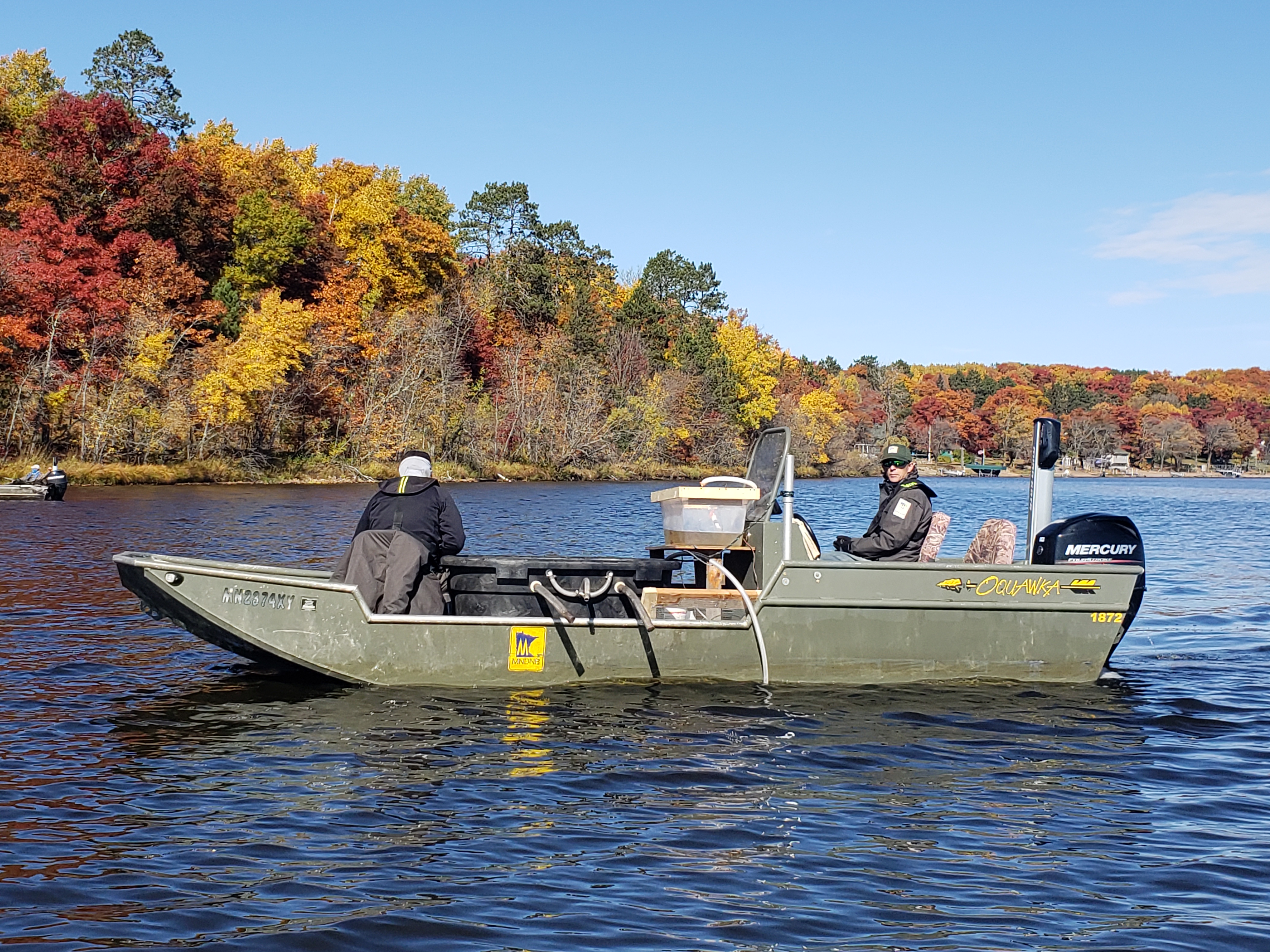Two people on a boat on the water with colorful fall trees in the background