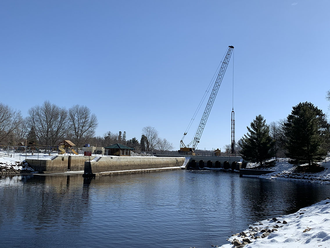 A body of water with a dam and a construction crane in the background. There is snow on the gorund.